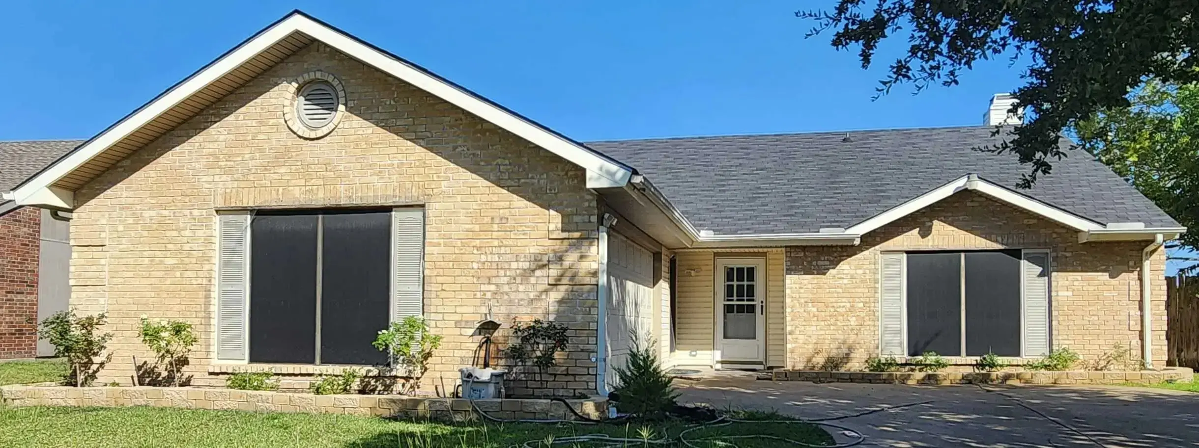 <p>A charming single-story brick ranch house is transformed with a cozy yet modern design. The house features large windows that enhance natural light, sleek wooden shutters, and built-in flower boxes beneath the windows. The front entry is updated with a contemporary energy-efficient door, surrounded by minimalist landscaping filled with easy-care native plants. The old driveway has been replaced with an eco-conscious permeable paving system, allowing for better water management. The distinctive gable roofs are highlighted with chic siding and a freshly applied coat of paint.</p>