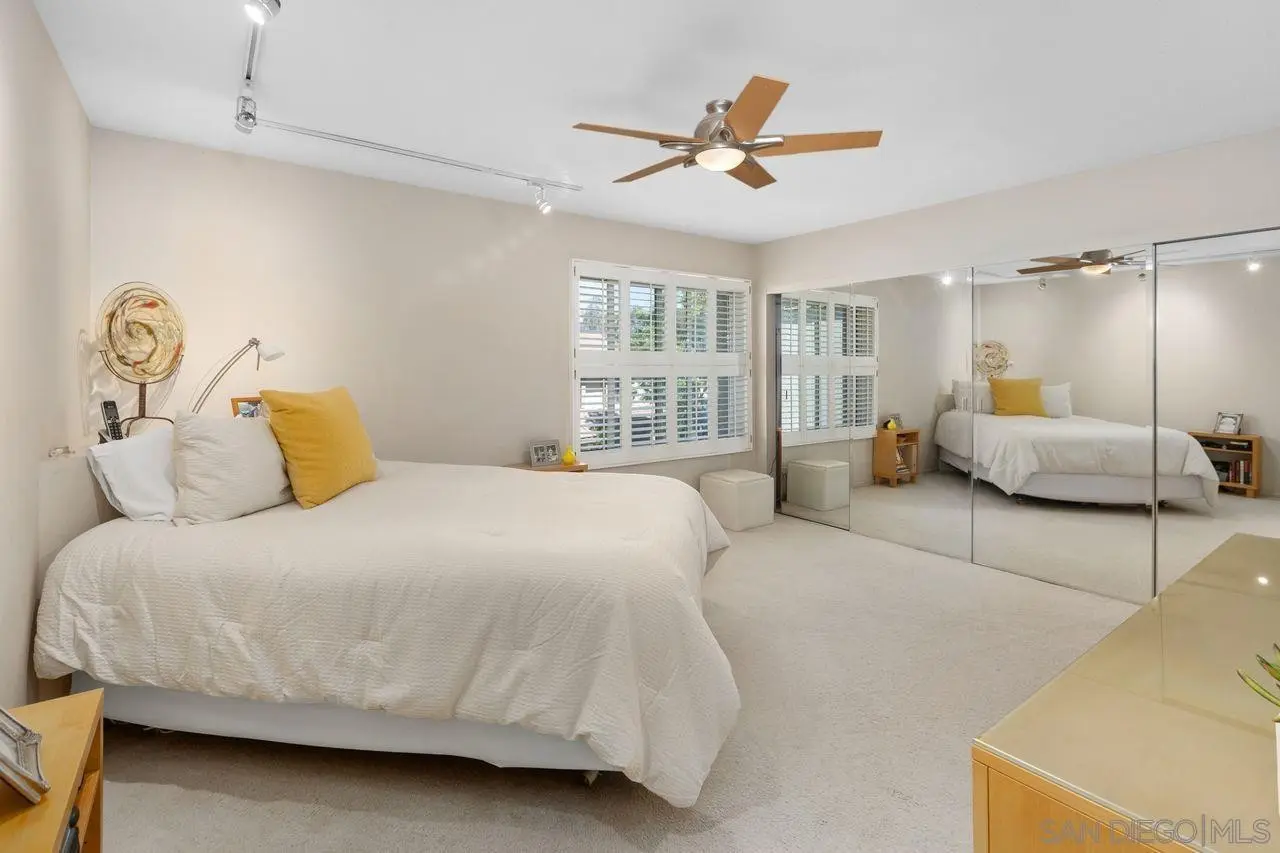 <p>A contemporary bedroom interior of around 15x15 feet. This room includes a queen-sized bed at the center. To its left, there's a sleek wooden desk with a minimalist aesthetic. On the right side of the bed, there's a compact vanity set with a mirror. The color scheme of the room involves neutral, warm hues, with concealed storage areas located underneath the bed and integrated into a sophisticated wall unit. The room is illuminated by recessed lights complemented by a ceiling fan, and the floor has a light wood finish. Abundant natural light permeates through large windows equipped with shutter blinds.</p>