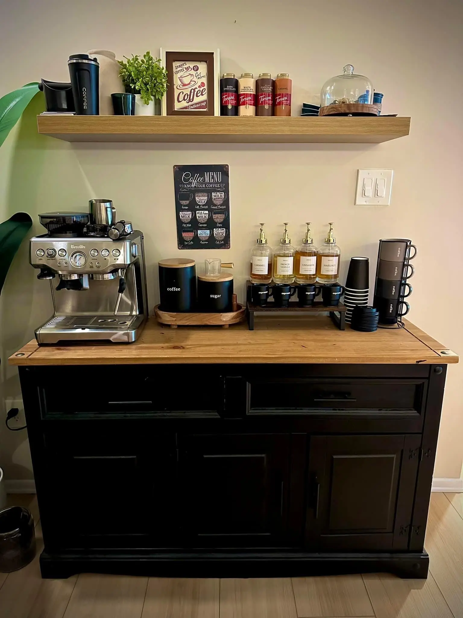 <p>Design a contemporary kitchen with a modern coffee station. The station features a sleek espresso machine resting on a marble countertop that is white in color. An integrated wine cooler sits beneath the countertop. The shelves, constructed with a light oak finish, hold neatly arranged coffee accessories. The backsplash is adorned with geometric tiles, giving it a stylish look. The kitchen, measuring approximately 6 feet wide and 3 feet high, has an ambient light fixture above the coffee station, along with additional storage units designed in a minimalist style for coffee pods and mugs.</p>