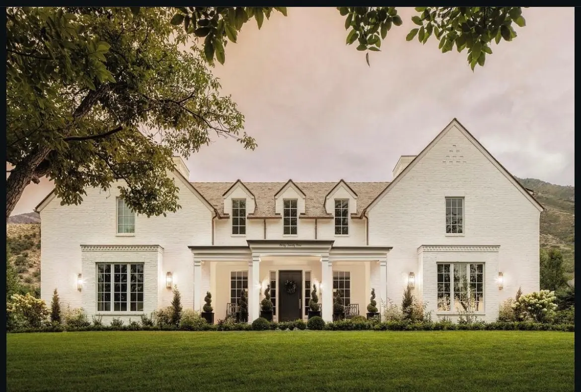 <p>Create an image of a traditional two-story house exterior finished in white brick, with symmetrical windows and a central entrance. The house is set against a backdrop of a lush, green front yard, with mature hedges and trees. The floor plan of the house contains a kitchen that has a sink overlooking a backyard, which connects to a dining area that opens up into a living room. The living room boasts a fireplace being flanked by built-in cabinets. The plan also shows a laundry room with vast folding space, a visually hidden mudroom with cabinets resembling lockers, and a kitchen equipped with a gas stovetop. There are in total four bedrooms and a study room located on the ground floor, with a bonus space available upstairs for future expansion possibilities. The house has a 3-car garage, including amenities like generator hookups, a basic dog wash area, and additional deep space for storage. The master suite includes an enclosed toilet area, a Roman-style zero-entry shower, and a loop leading to the laundry. Lastly, all the renovations applied adhere to a costing budget of $5,000, and a remodelling duration of over 6 months.</p>