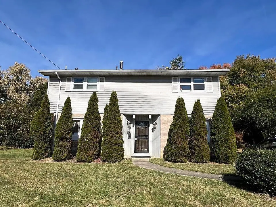 <p>A two-story suburban residence with a contemporary exterior makeover. The house measures roughly 25 feet in width and 20 feet in height. The old vinyl siding has been replaced with a sleek, dark gray rendering to exude a modern ambiance. Large, energy-conserving windows with black frames are installed on both floors for more natural light. A minimalistic wooden door with a frosted glass section marks the entrance. The pathway has been revamped with charcoal gray stepping stones leading to the front door. The landscape showcases well-groomed, symmetrical conifer trees aligned uniformly by the house, amidst fresh green grass. Contemporary outdoor lighting fixtures have been mounted near the entrance for illumination.</p>