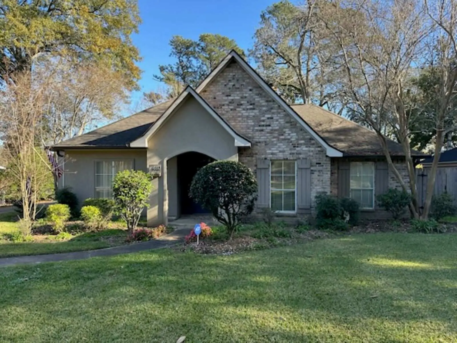 <p>Generate a highly realistic image of a renovated residential exterior. The house features a stone pathway leading to the entrance, encircled by an array of vibrant flowers and shrubs measured approximately 10 by 15 feet. On both sides of the door, there are modern outdoor lighting fixtures. A small sitting zone with a bench is visible. Even though the house maintains its original structure and rooftop, it has received a new coat of warm, inviting paint. The enhanced landscape includes manicured trees and abundant verdant grass.</p>