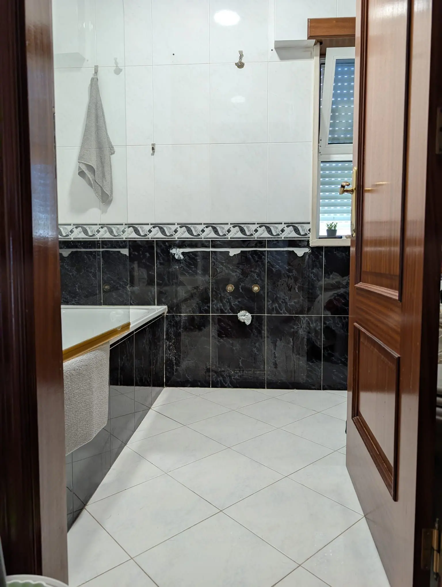 <p>Generate a highly realistic image of a remodeled bathroom, measuring approximately 8 feet by 6 feet. The design should incorporate a half-wall adorned with dark marble tiles, juxtaposed with light-colored tiles on the remaining walls. Existing features like the towel hook, wooden door, small shelf, and window should be retained. Add a stylish mobile with geometric shapes, artistically hanging above the bathtub area. This addition should infuse a modern design element while harmoniously blending with the existing style of the bathroom.</p>